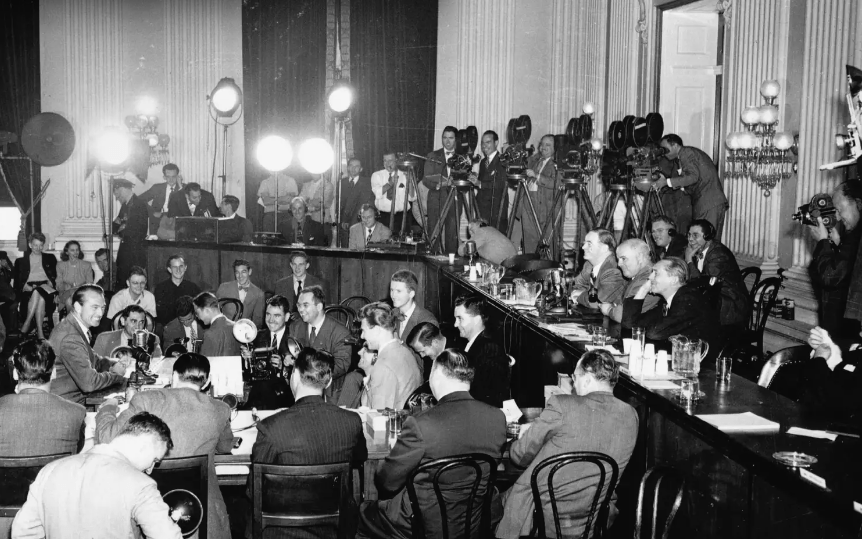 This black and white photograph depicts a HUAC hearing in the 1950s. The committee members are seated at a long table, facing witnesses who are testifying. The photograph captures the seriousness and tension of the hearings, which were often used to investigate alleged communist activity in the United States.